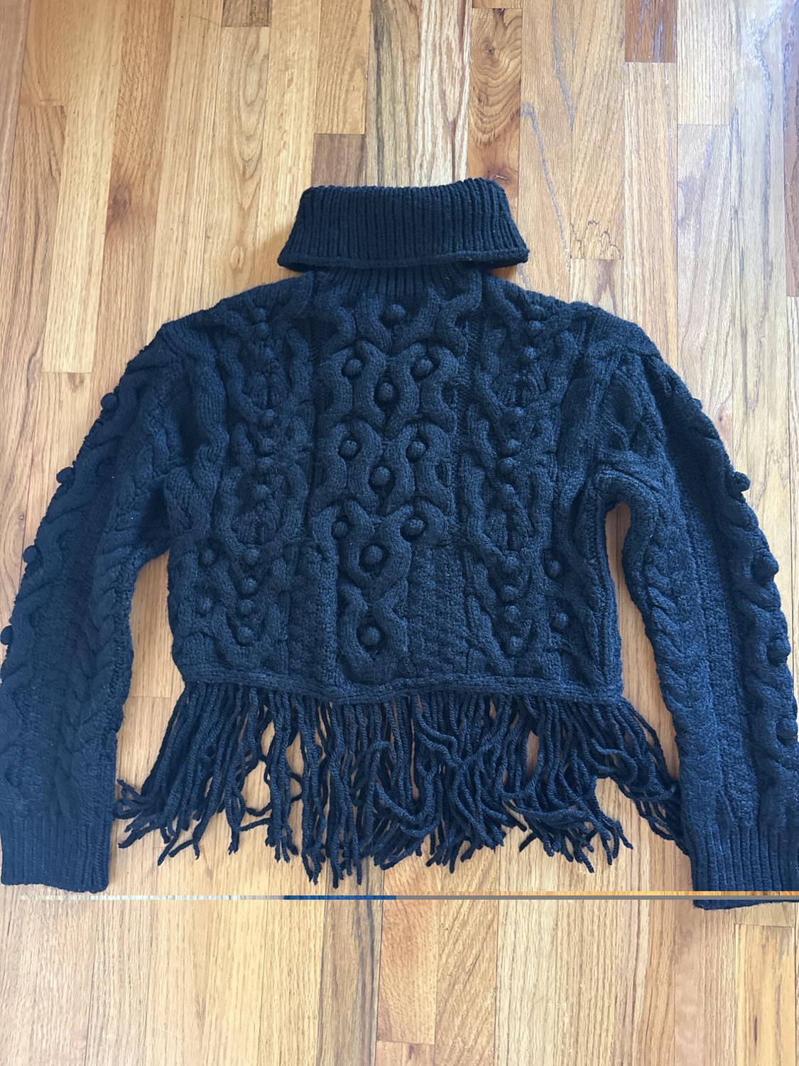 Black chunky knit roll neck jumper with a cable knit design and fringing on the bottom hem, on a light wooden floor.