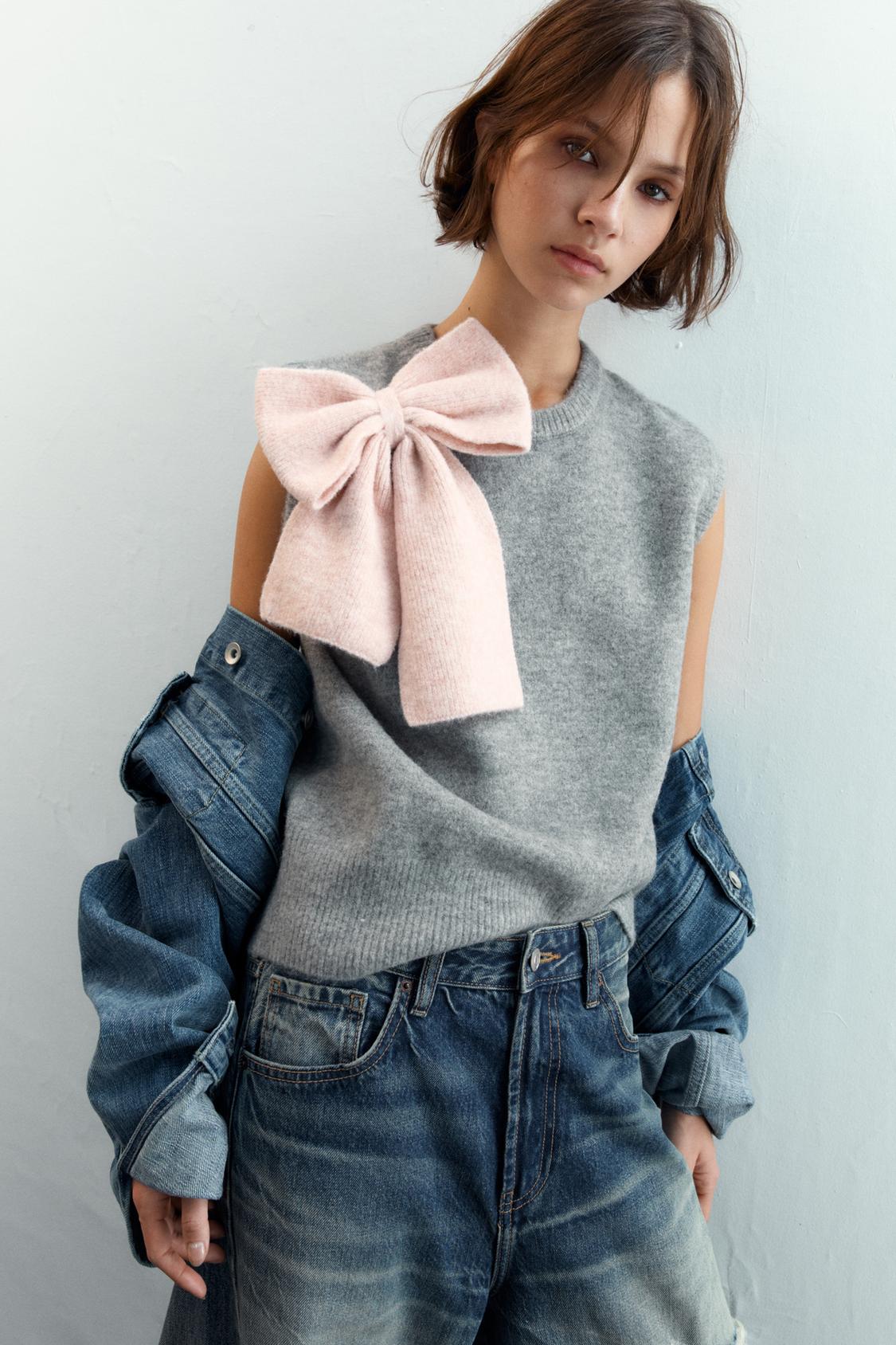 Siv brezrokavnik z rožnato pentljo na rami, modra denim jakna in obrabljene kavbojke.