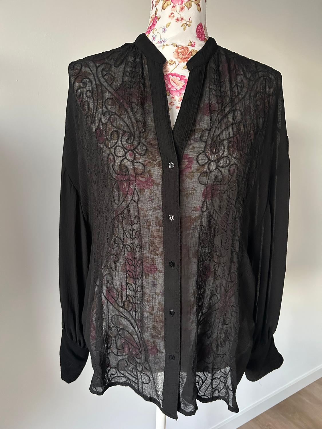 Sheer black blouse with floral embroidery on a mannequin with a floral print. White background and wooden floor.