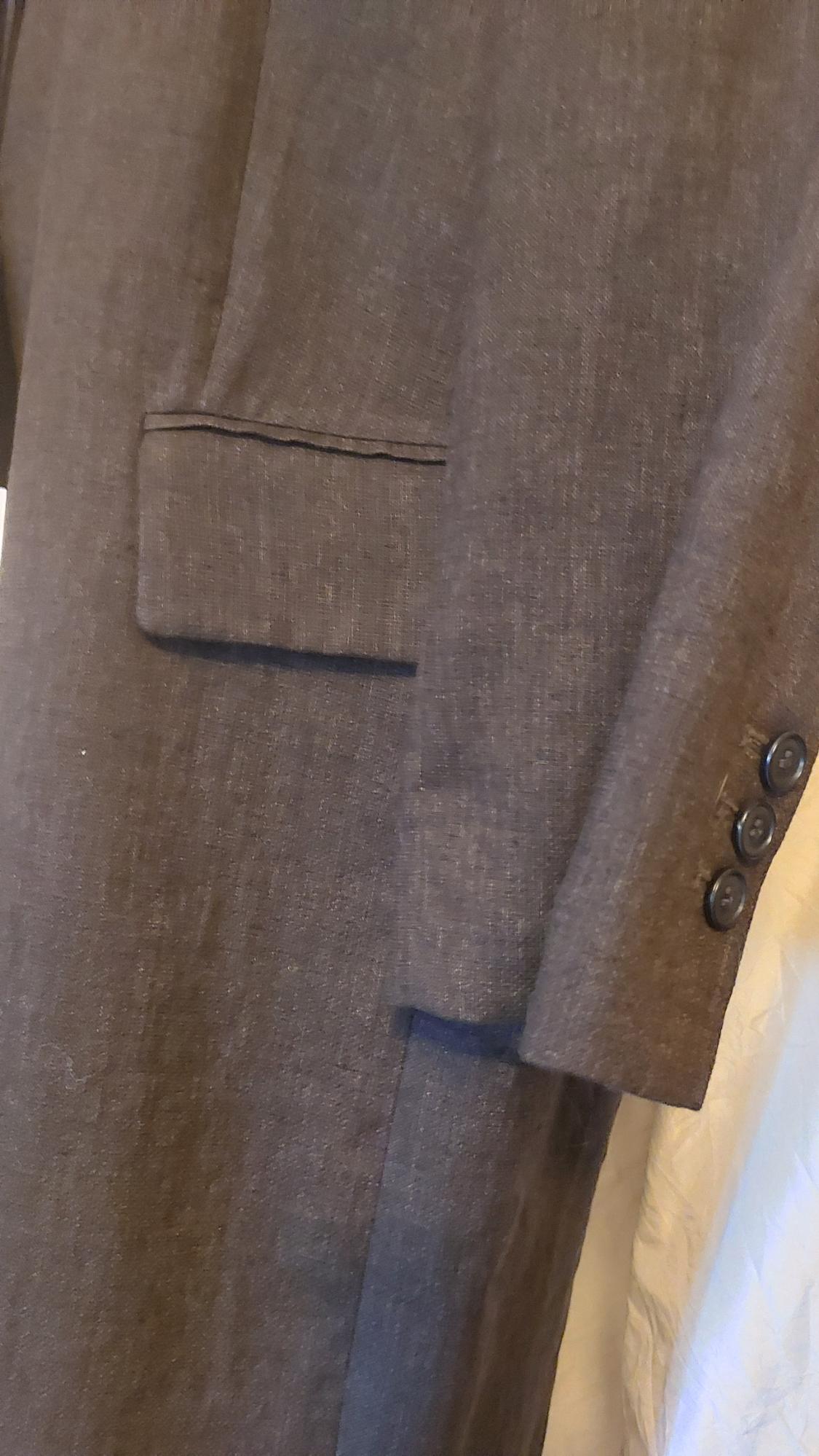 Detail of a brown jacket with visible texture, showing a pocket and three buttons on the sleeve against a light background.