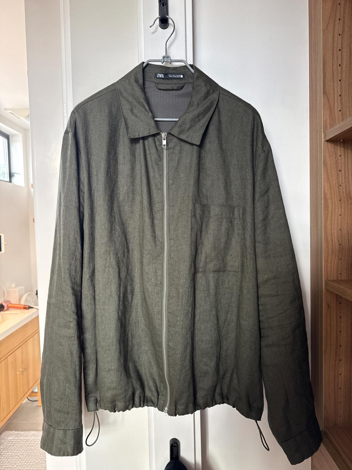 Olive green jacket with a zip, hanging on a hanger inside a white wardrobe. Visible label with text "ZARA".