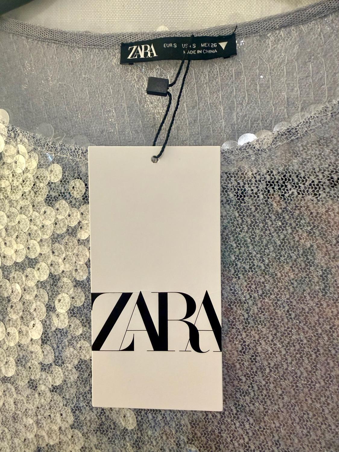 Grey garment with sequins. Black label with the text ZARA EUR S USA S MEX 26 MADE IN CHINA. White label with the text ZARA.