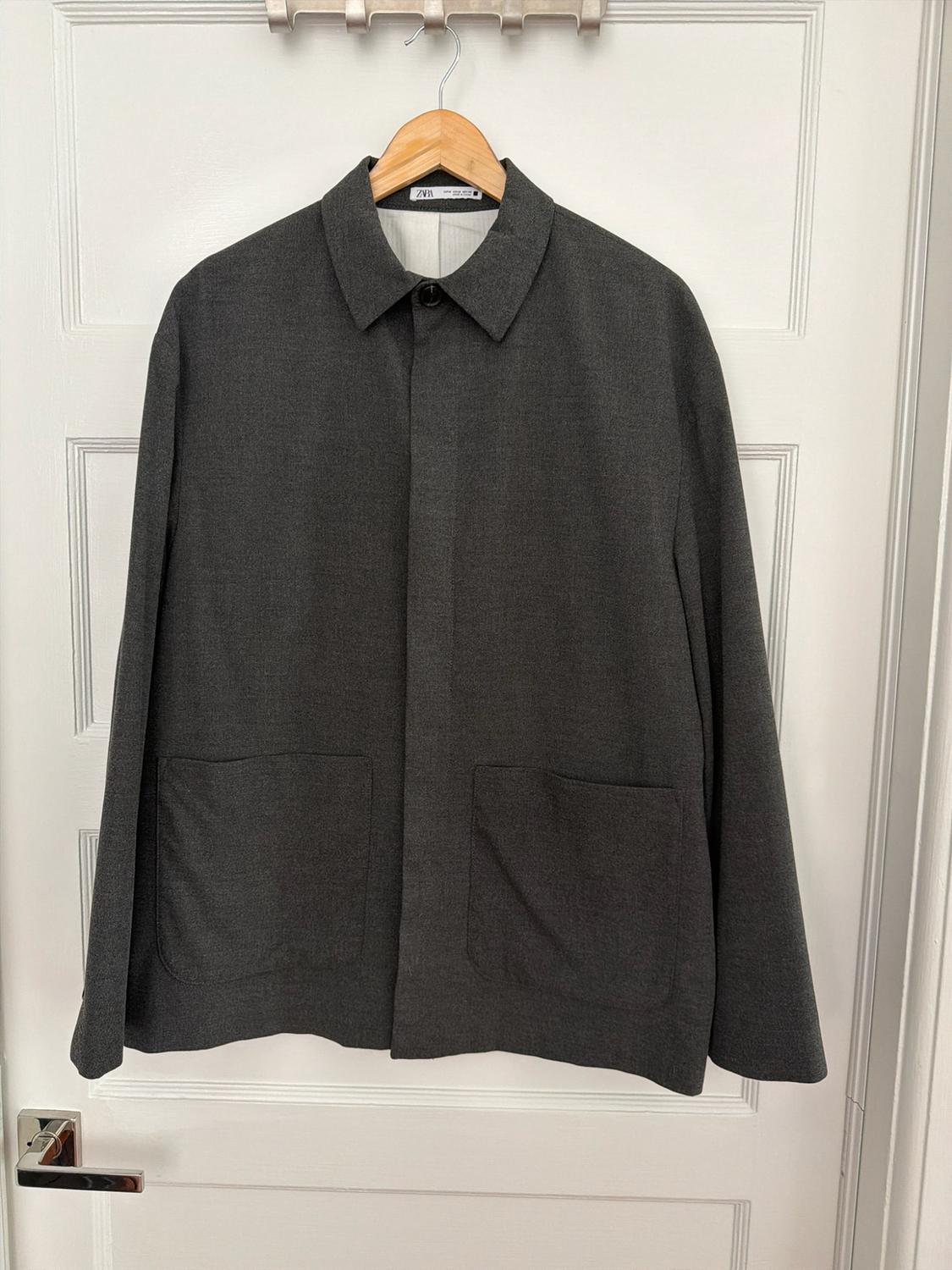 Dark gray jacket with a collar and patch pockets, hanging on a wooden hanger over a white door. Label with the text Zara.