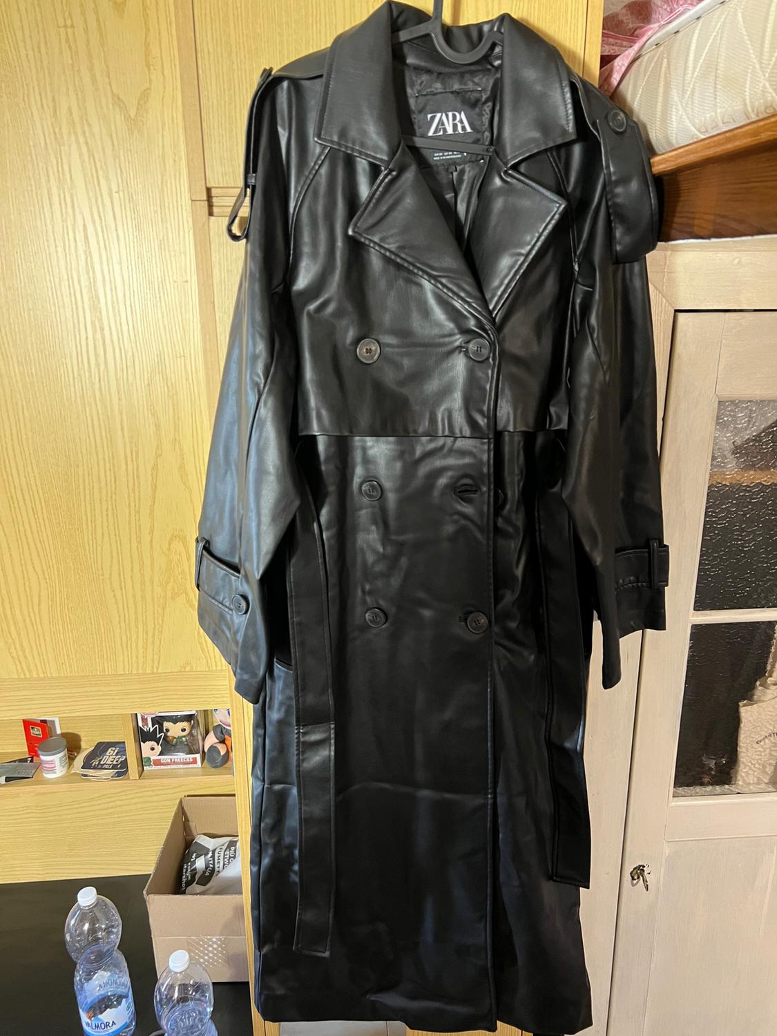 Long black trench coat with belt hanging in a wooden wardrobe. Label with ZARA text. Casual setting with bottles and boxes.