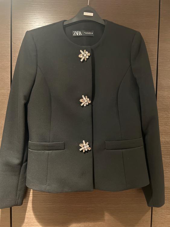 Black, long-sleeved jacket with floral rhinestone brooches, hanging on a black hanger with the "ZARA" logo visible.