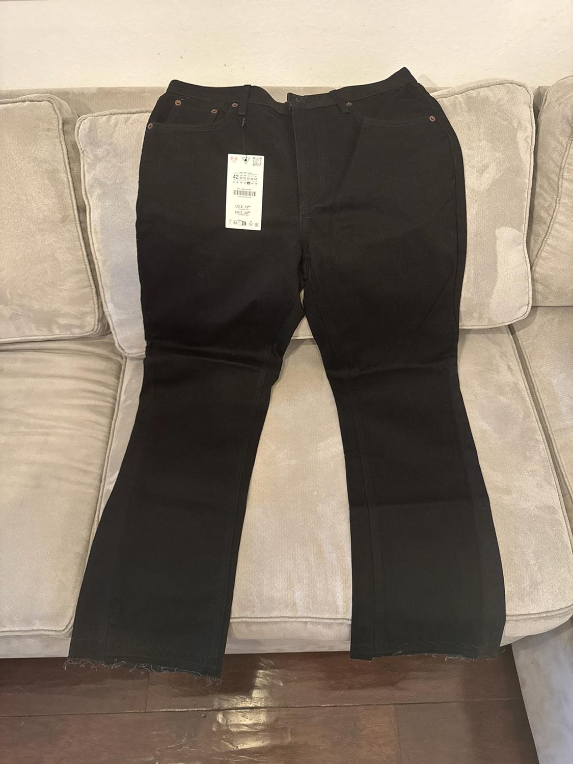 Black pants with a tag laid out on a beige sofa. Straight-leg design
with a frayed hem and visible wooden floor.