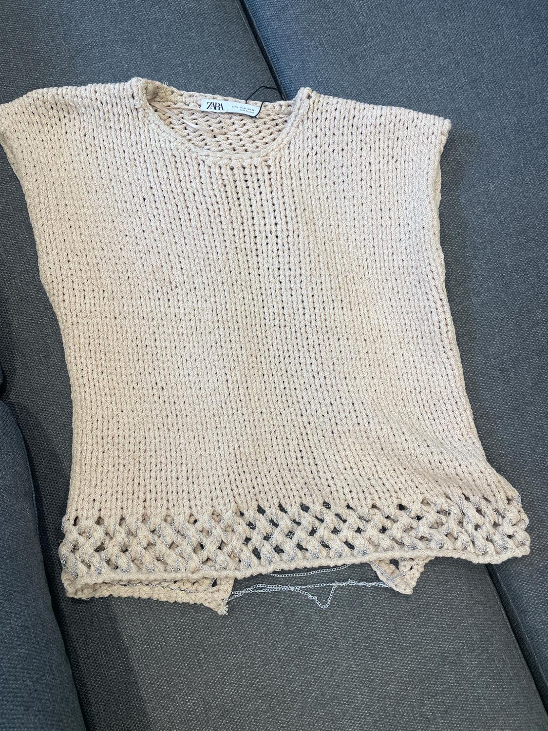 Beige knitted waistcoat with an openwork design on the bottom edge, on a grey sofa. Visible brand label.