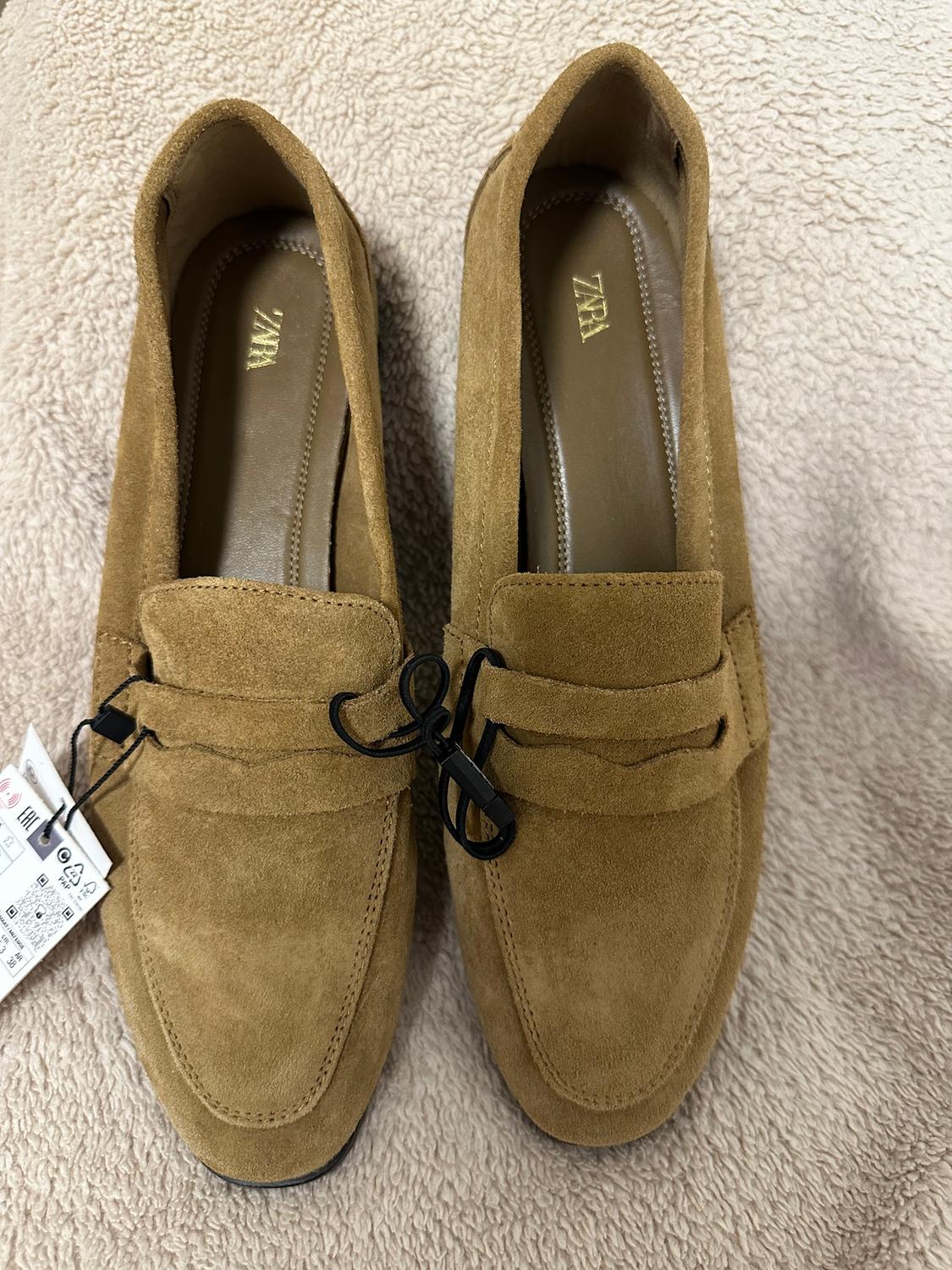 Brown suede shoes with a black bow and visible tag, on a beige fabric background. ZARA brand on the insole.