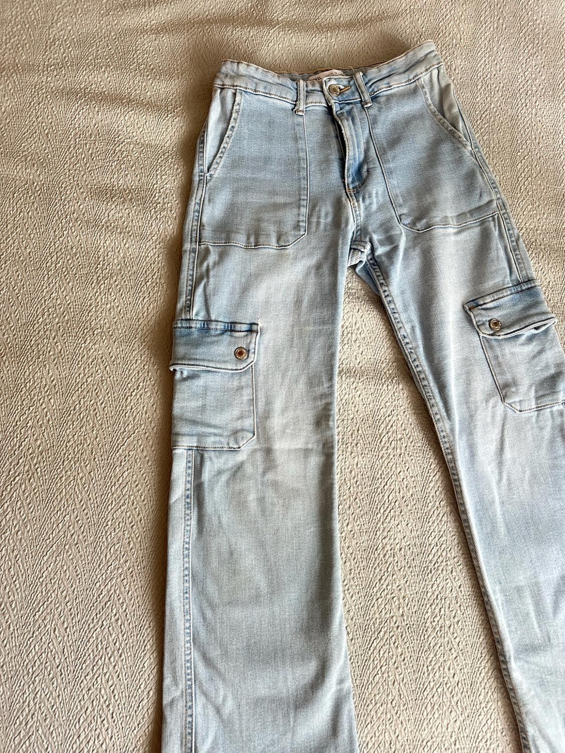 Light blue cargo jeans with front and side pockets, against a textured beige fabric background.