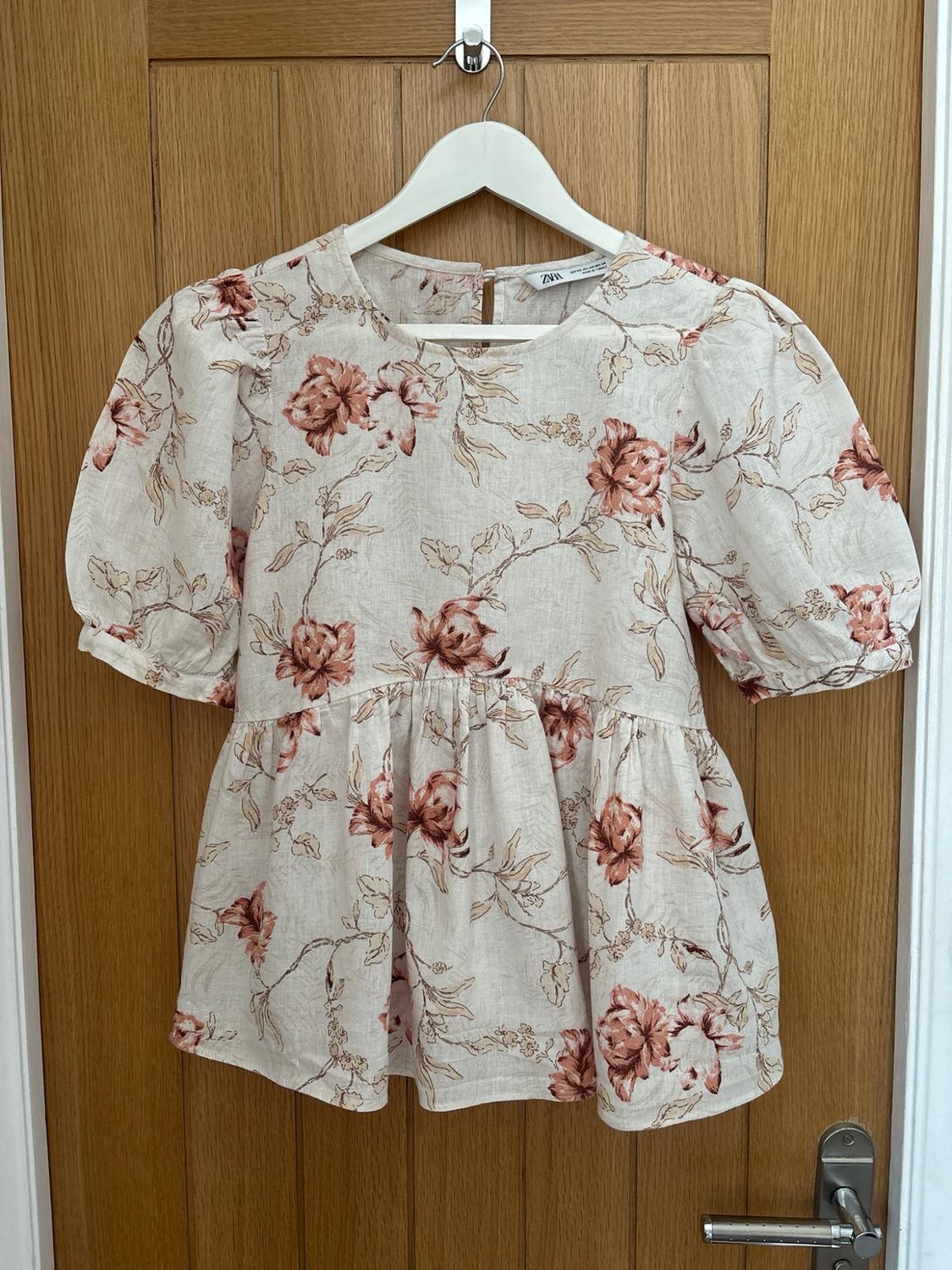 Light-coloured blouse with a pink floral print, short puffed sleeves and a flared hem, hanging on a wooden door. Label with the text ZARA.