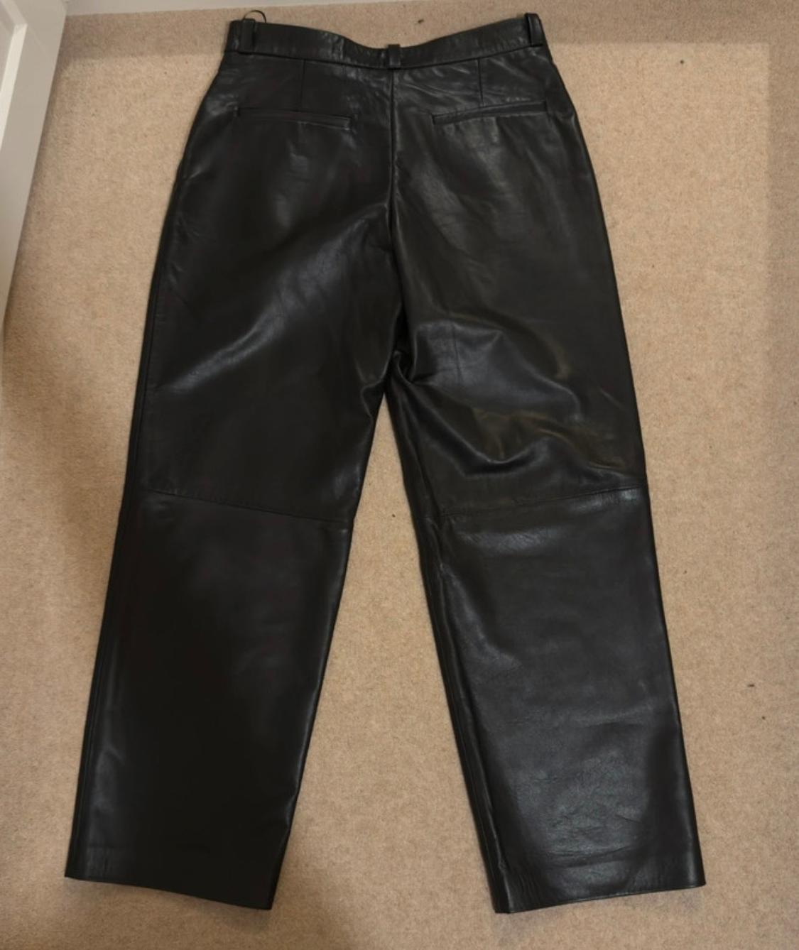 Black leather trousers laid flat on a beige rug, showing back pockets and a waistband with belt loops.