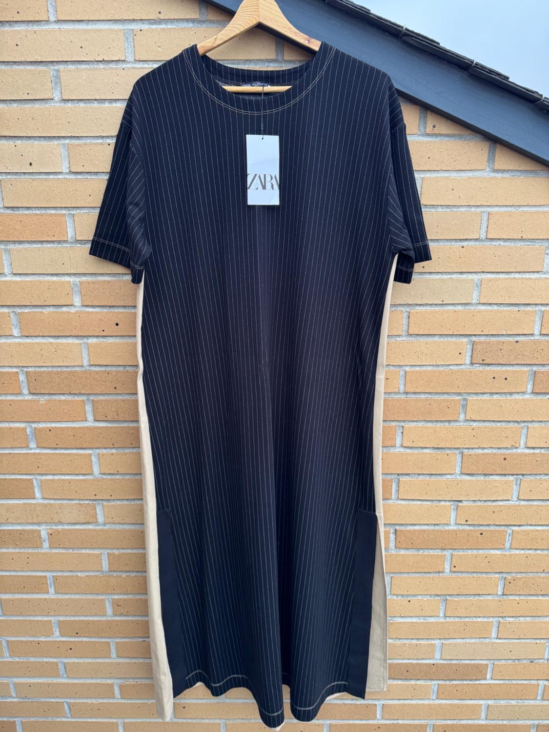 Black dress with vertical stripes hanging on a hanger in front of a brick wall. Visible label with the text "ZARA".