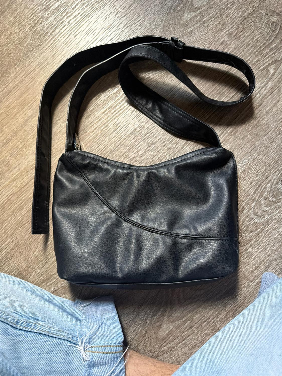 Black shoulder bag with stitching detail on a wooden floor. At the bottom, legs in ripped blue jeans peek out.