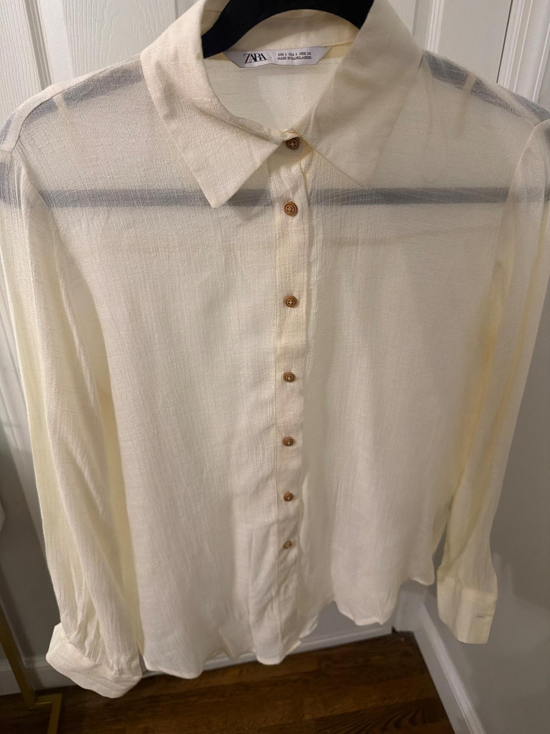 Semi-sheer white long-sleeved shirt with brown buttons, hanging on a black hanger in front of a white door.