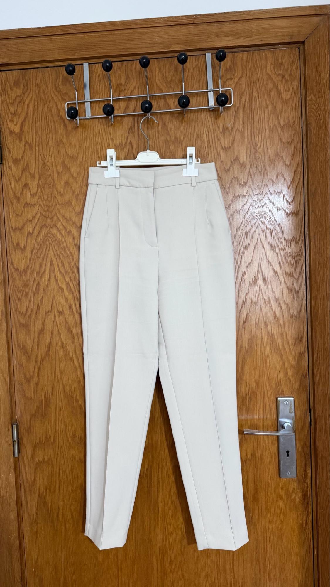  dress trousers with a front crease, hanging on a white hanger on a wooden door with a metal coat rack.