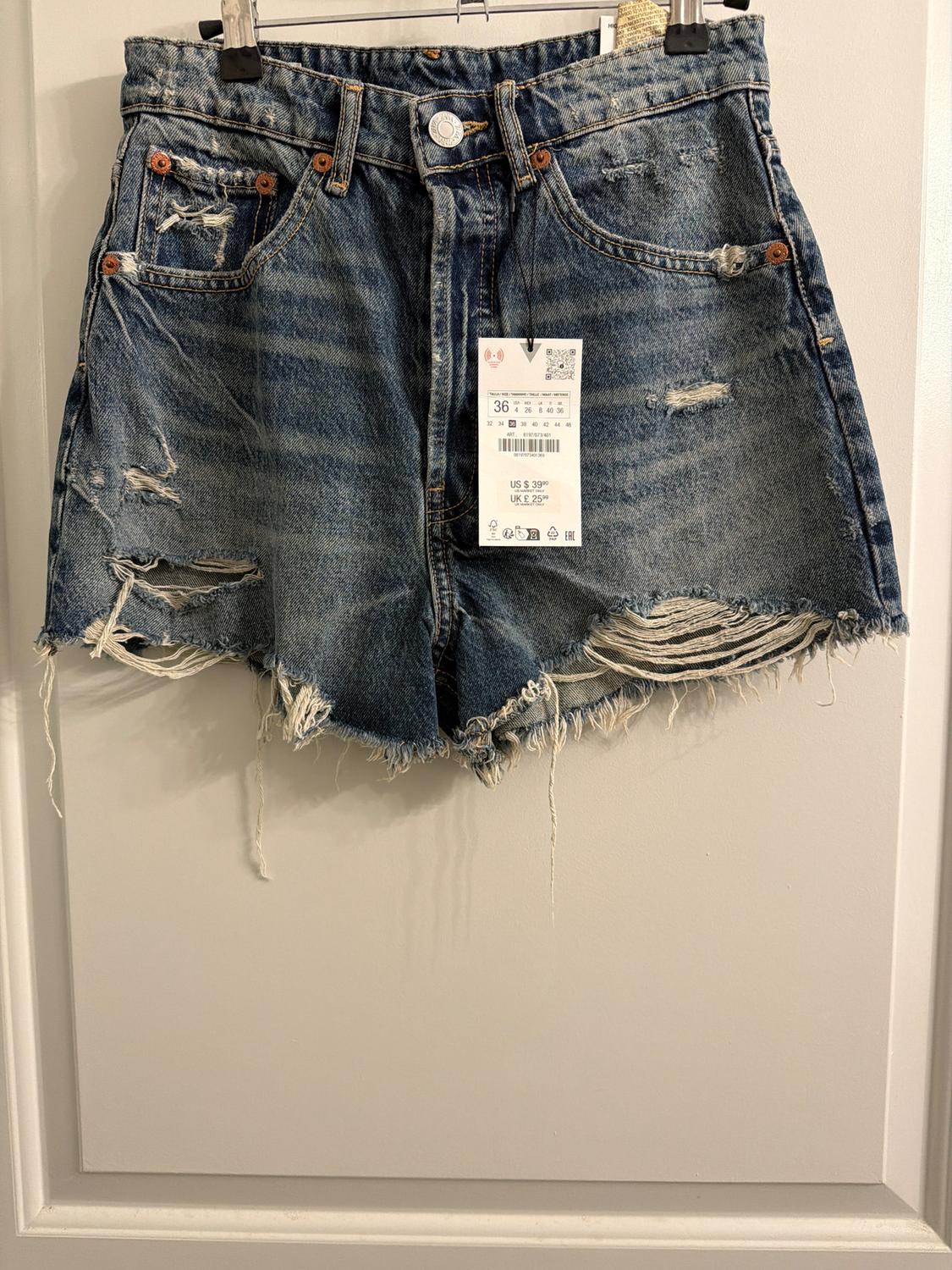 Distressed denim shorts with rips and a hangtag. They are hanging on a hanger against a white background.
