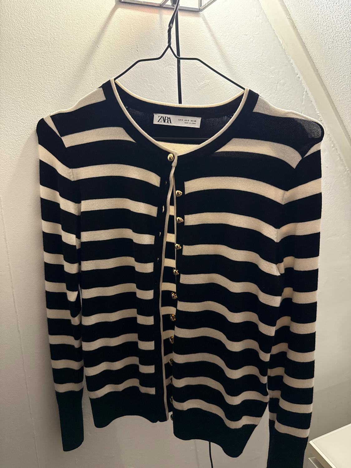 Black and beige striped cardigan hanging on a hanger. Visible label with text: "ZARA". Gold buttons on the front.