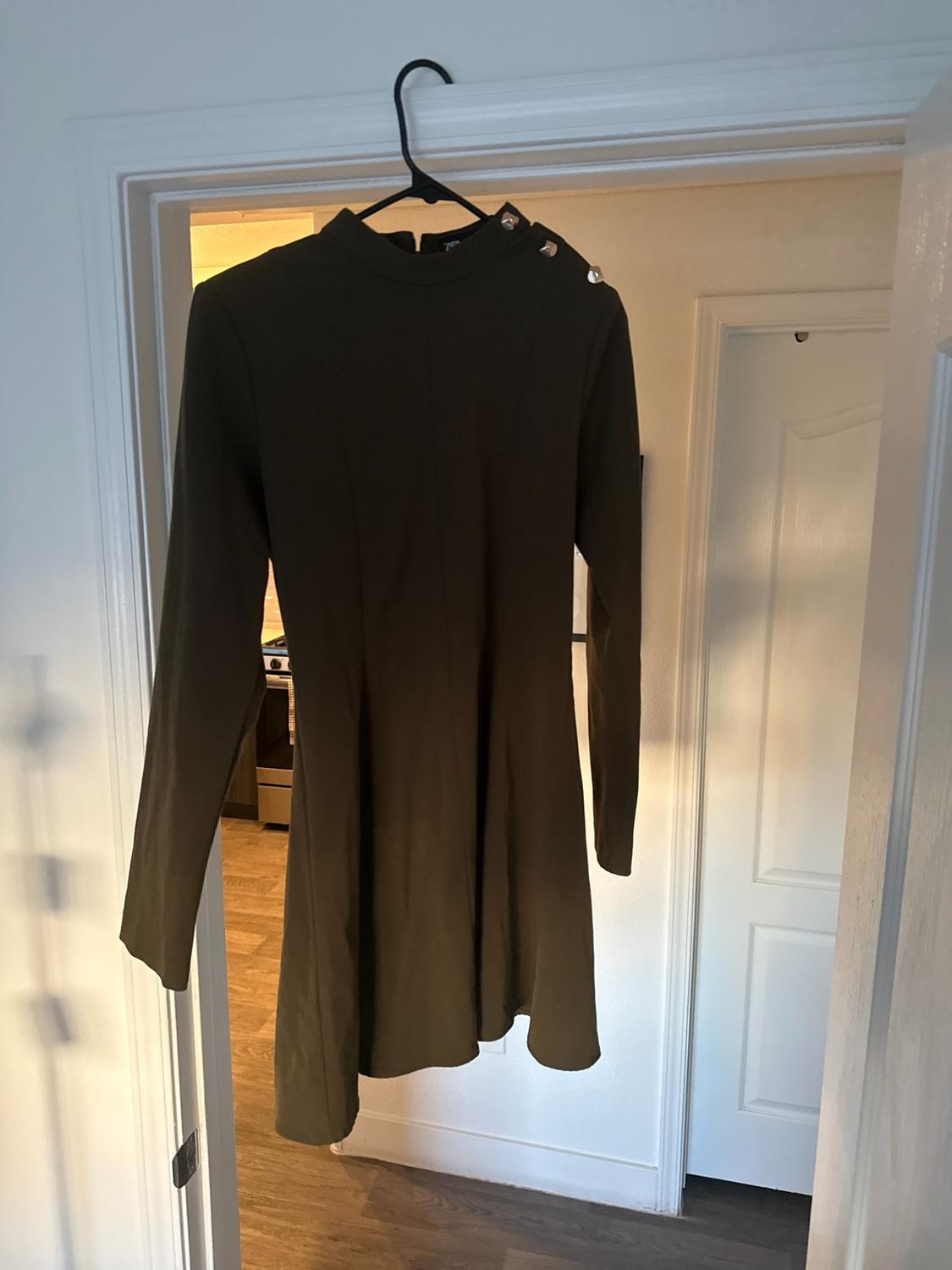 Olive-green short dress with long sleeves and a high neck with buttons on the shoulder, hanging on a hanger in the frame of a white door.