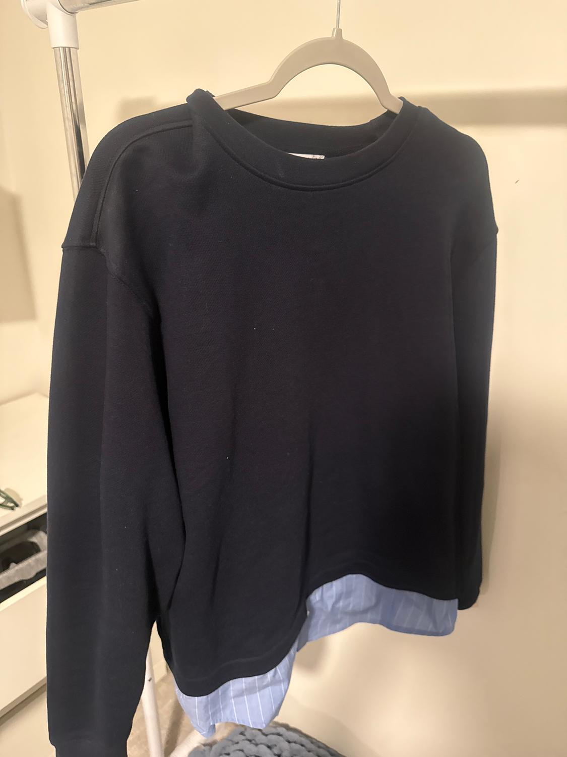 Black jumper hanging on a hanger, with a light blue striped shirt peeking out from underneath, against a beige background.