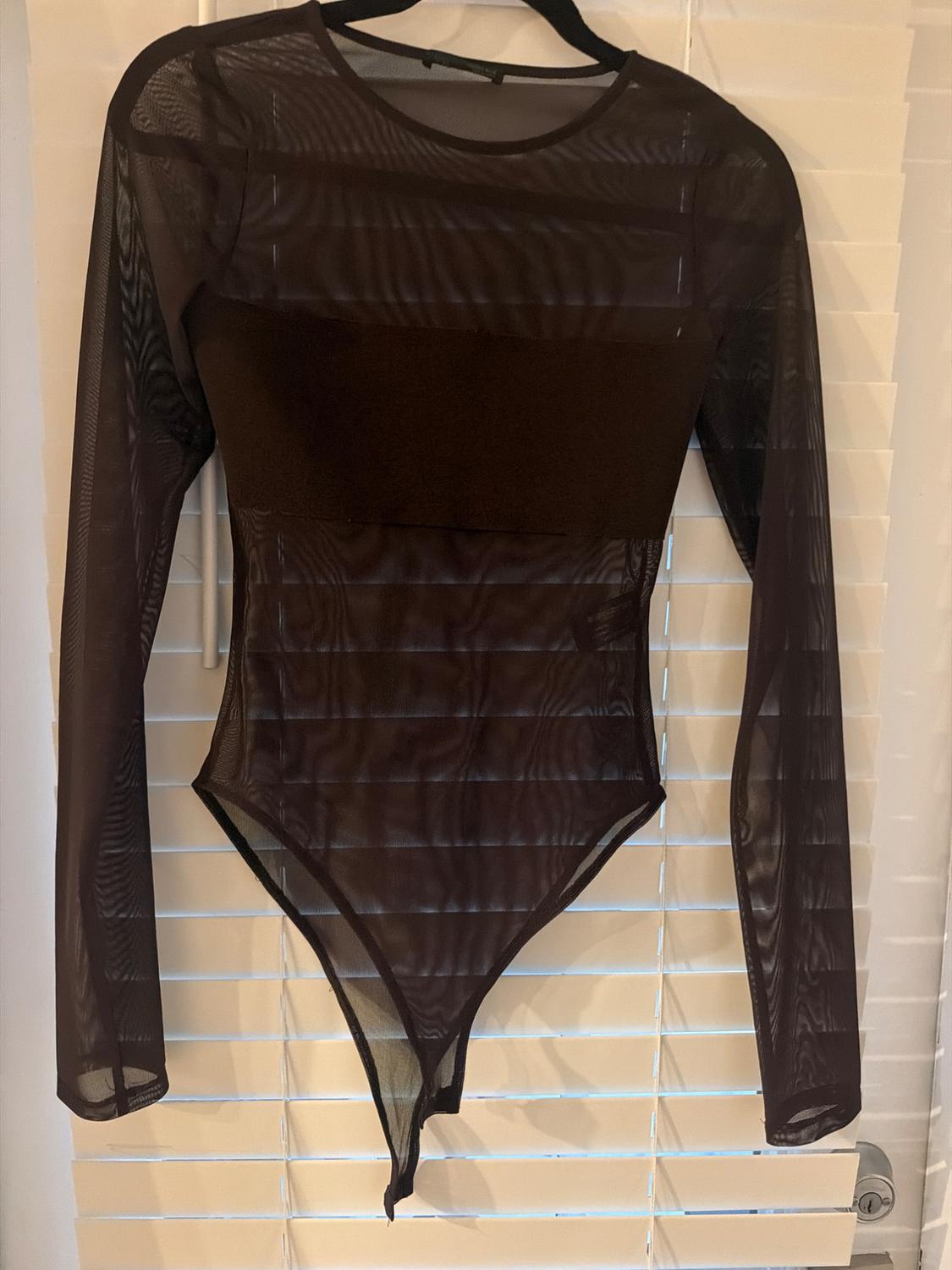 Long-sleeved black mesh semi-sheer bodysuit with an opaque panel on the chest, hanging on a hanger in front of white blinds.