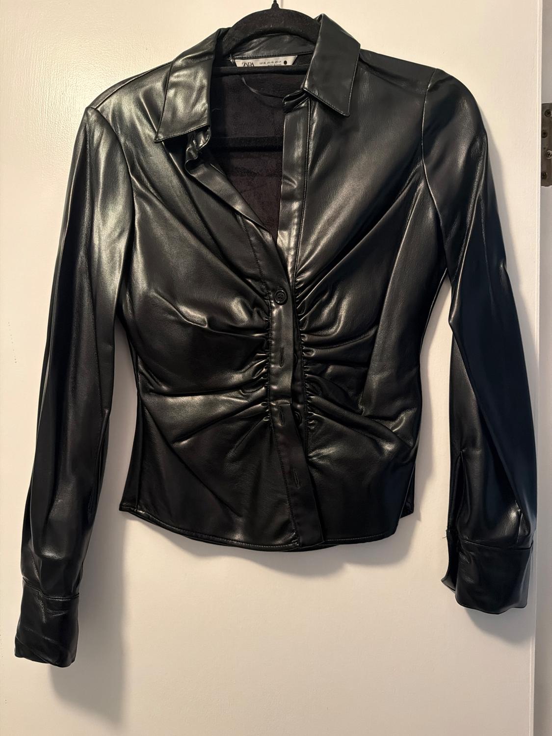 Black faux leather shirt with ruched detailing in the centre, hung on a hanger against a white background.