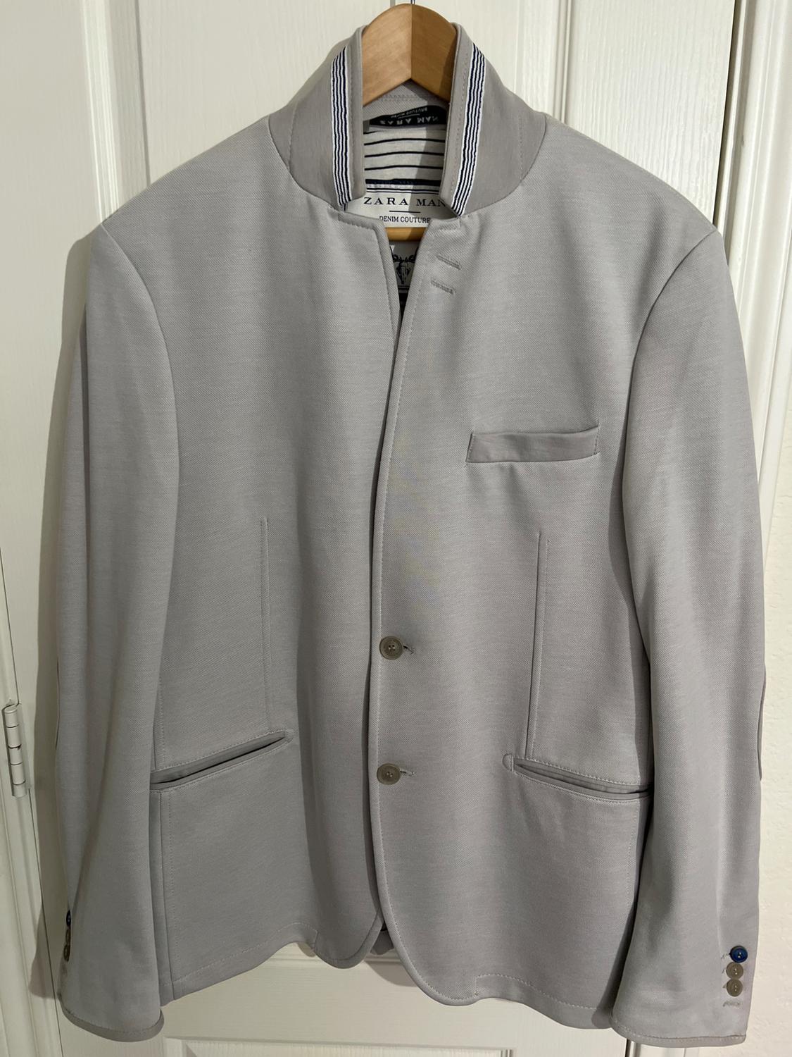 Light gray jacket hanging on a wooden hanger, with a mandarin collar and two visible buttons. Visible inner label: "ZARA MAN".