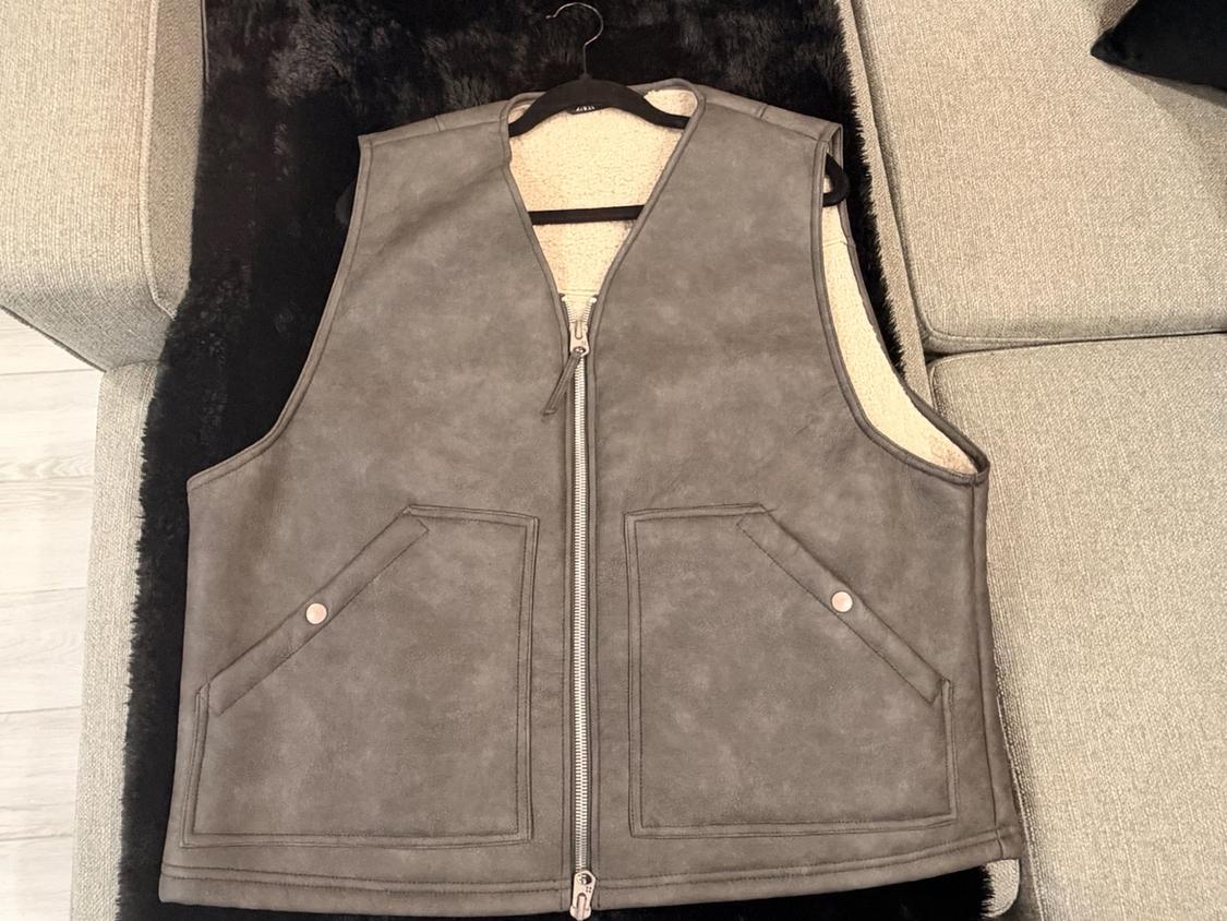 Gray zip-front vest with side pockets and a soft-textured inner lining, hanging on a hanger over a sofa.