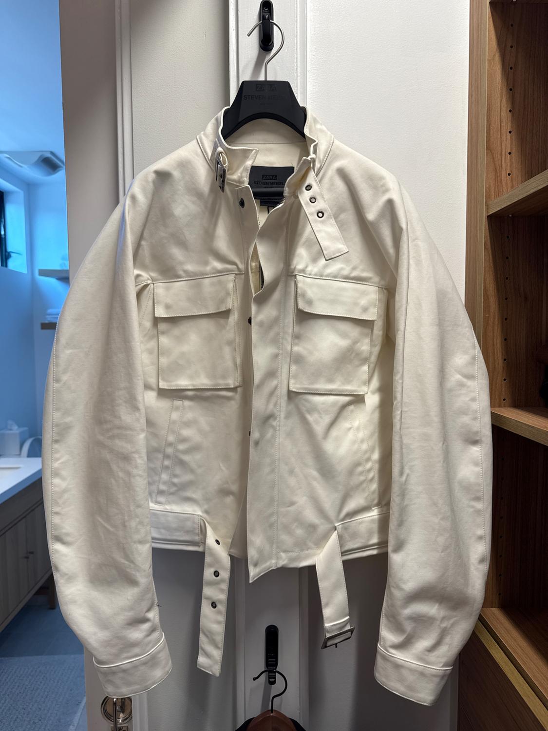 A white jacket hanging on a black hanger inside a wardrobe. It has front pockets and a waist belt.