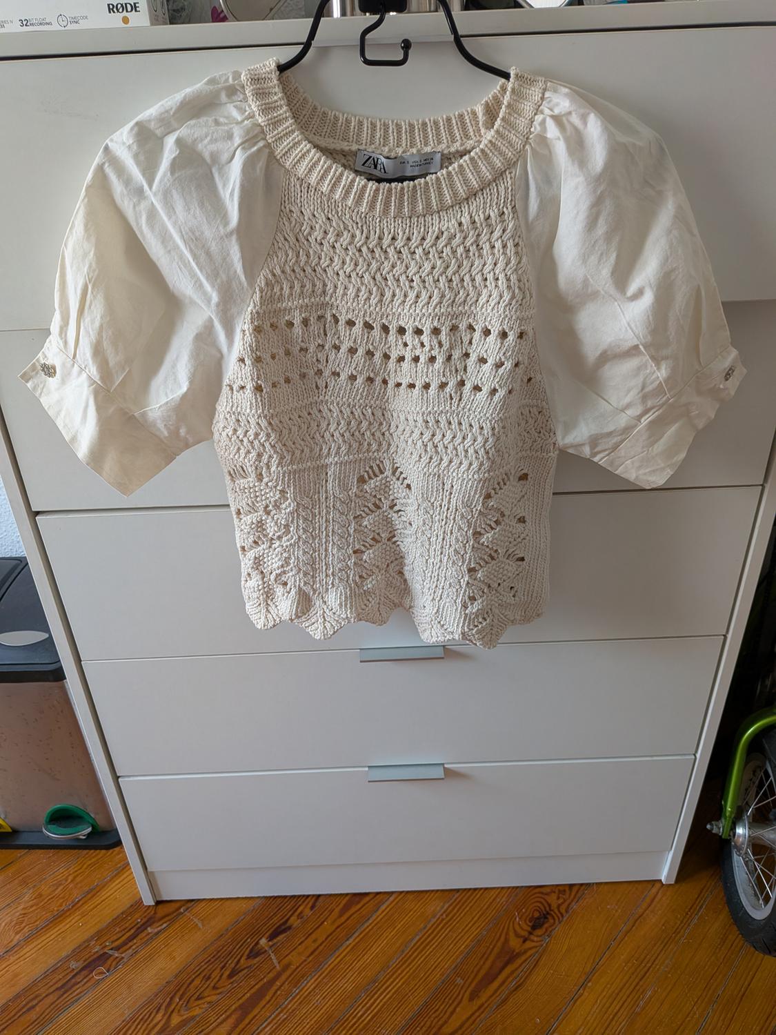Cream open-knit top with puff sleeves hanging on a hanger in front of a white chest of drawers. Visible text: ZARA, RØDE.