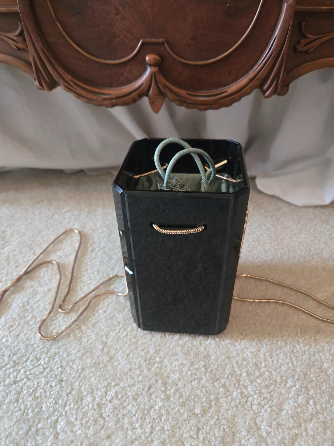 Black hexagonal handbag with a gold chain on a light-coloured rug, in front of a carved wooden cabinet.