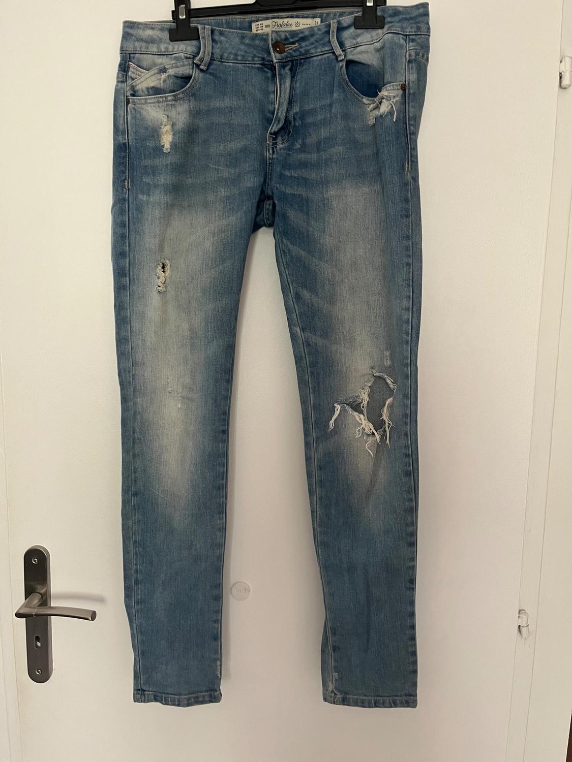 Front view of blue jeans with a distressed effect and rips on the right leg, visible front pockets.
