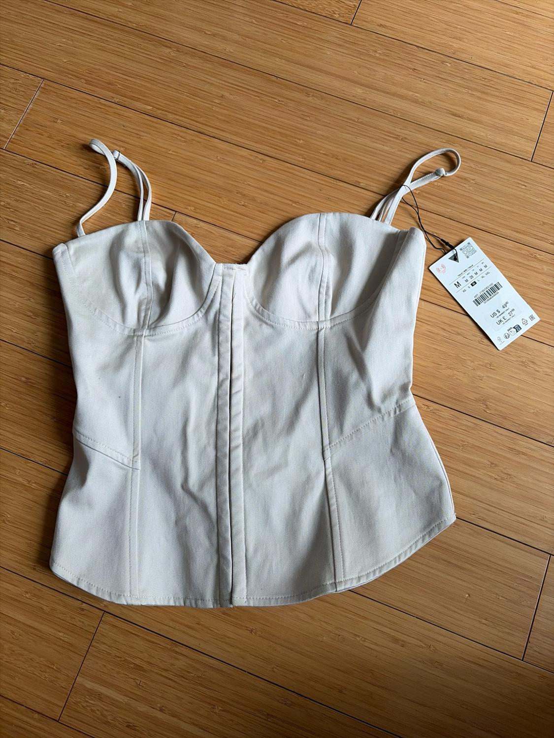 White corset top with thin straps on a wooden floor. Label text: M, US $ 49.90, UK £ 27.99.
