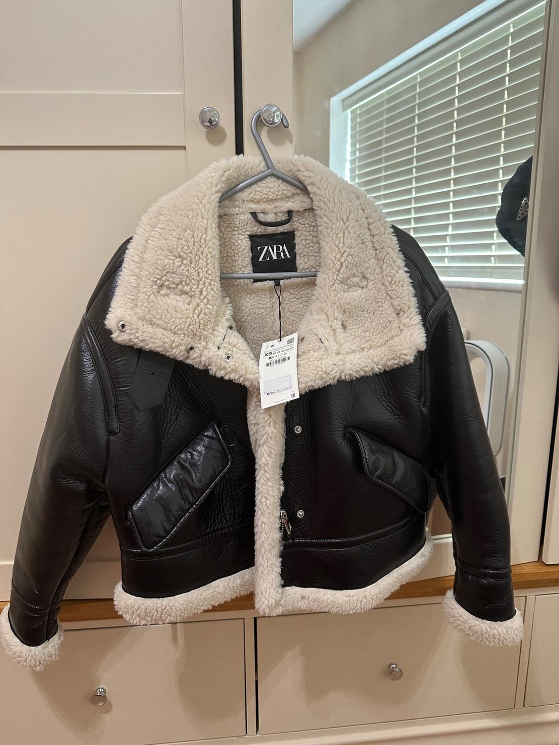 Black jacket with white shearling lining hanging on a hanger. Visible label with the text "ZARA". Beige wardrobe background.