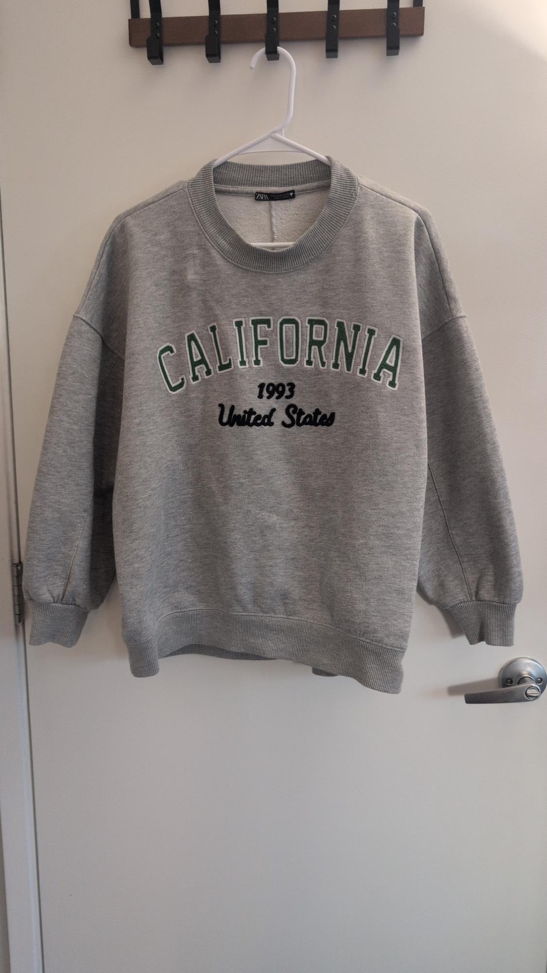 Gray crewneck sweatshirt hanging on a white door. Across the front, the text CALIFORNIA 1993 United States is printed.