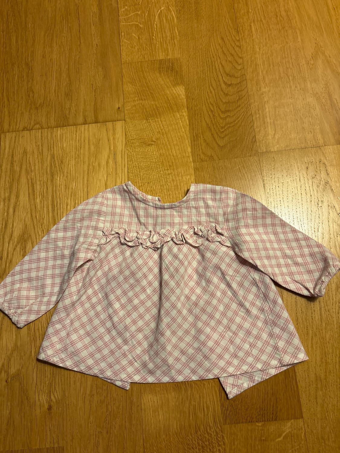 Long-sleeved children's blouse with pink and white checked print, ruffle detail on the chest, on a light wooden floor.