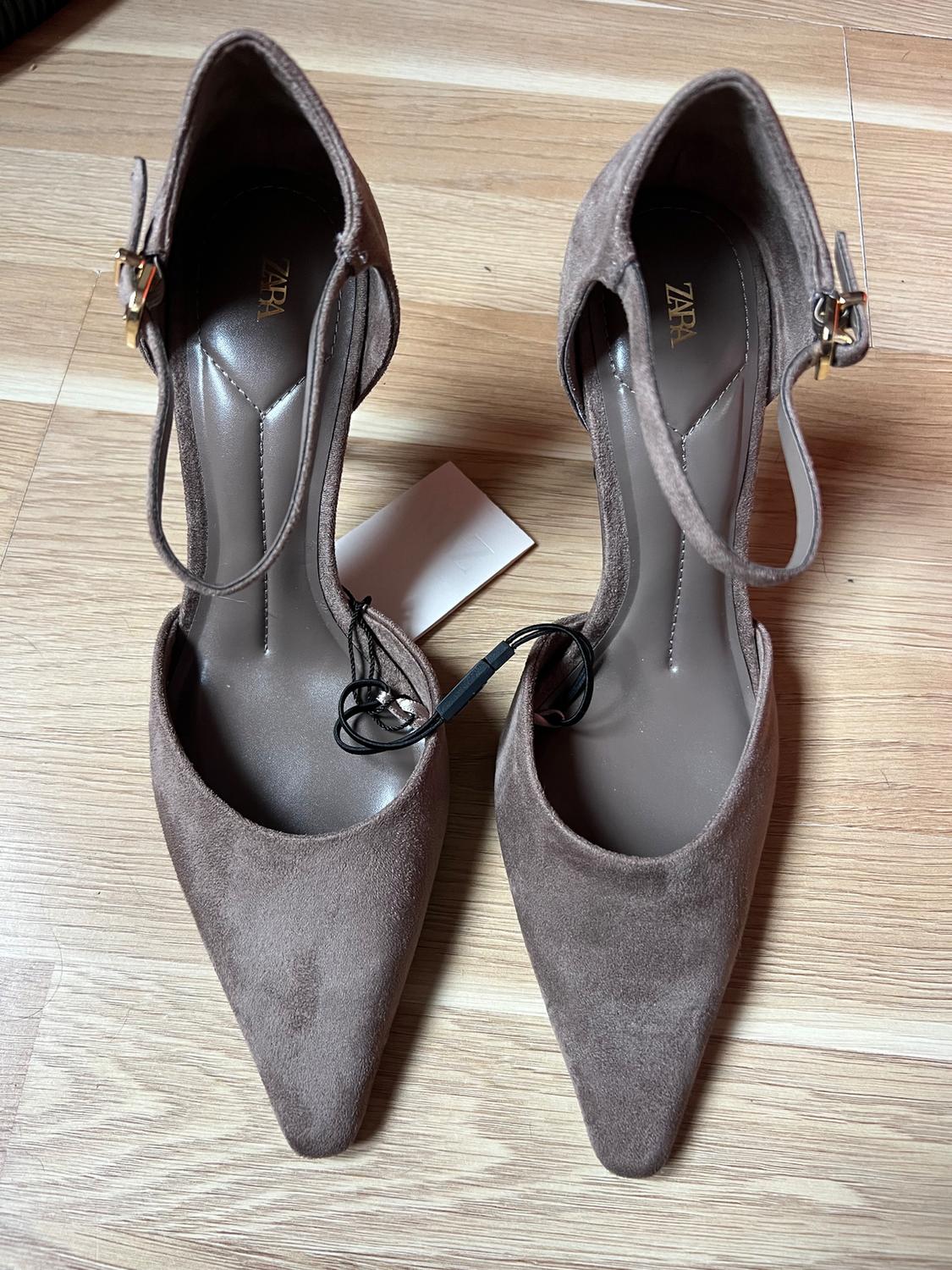 Brown stiletto heels with a pointed toe and ankle strap on a wooden floor. Visible label with text "ZARA".