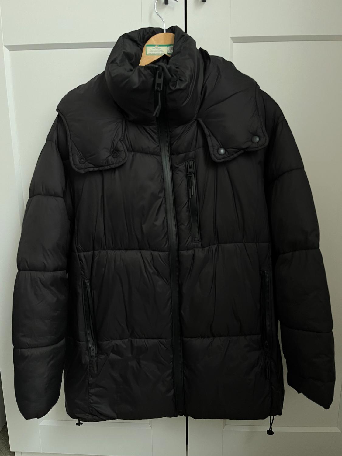 Black padded jacket hanging on a wooden hanger in front of a white wardrobe. Design with a high neck and zip fastening.