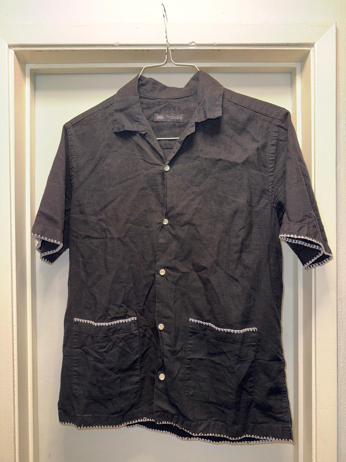 Black short-sleeved shirt with white buttons and two front pockets, hanging on a wire hanger on a white door.
