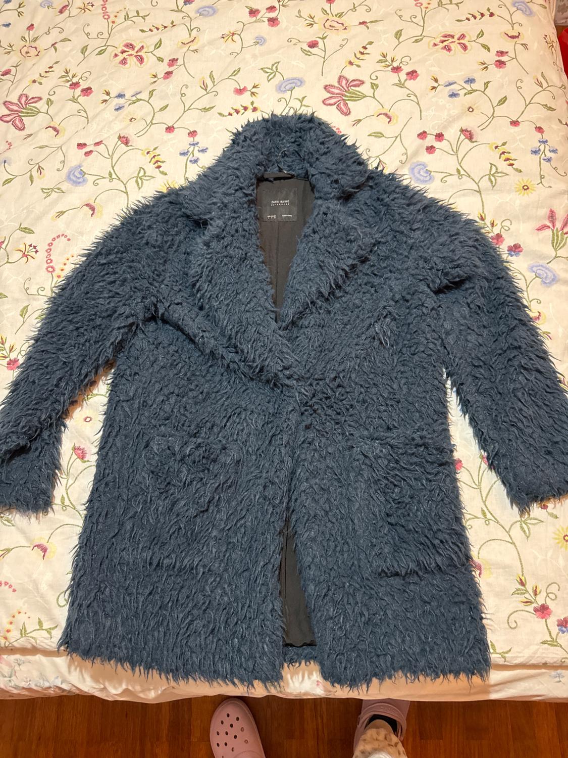 Σκούρο μπλε παλτό από γούνα με ετικέτα ZARA BASIC πάνω σε ένα κρεβάτι με ανθισμένα σεντόνια. Από κάτω προεξέχουν μερικά πόδια με ροζ τσόκαρα.