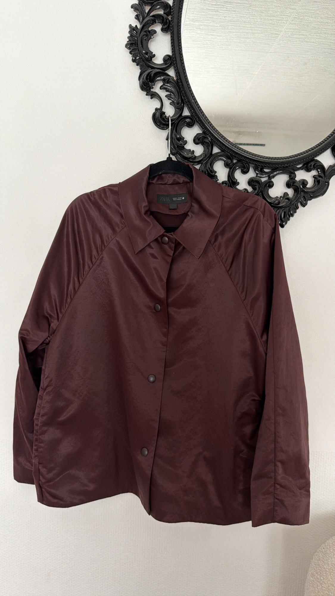 Burgundy button-up jacket hanging on a hanger under an ornate black mirror on a white wall. The inside label says ZARA.