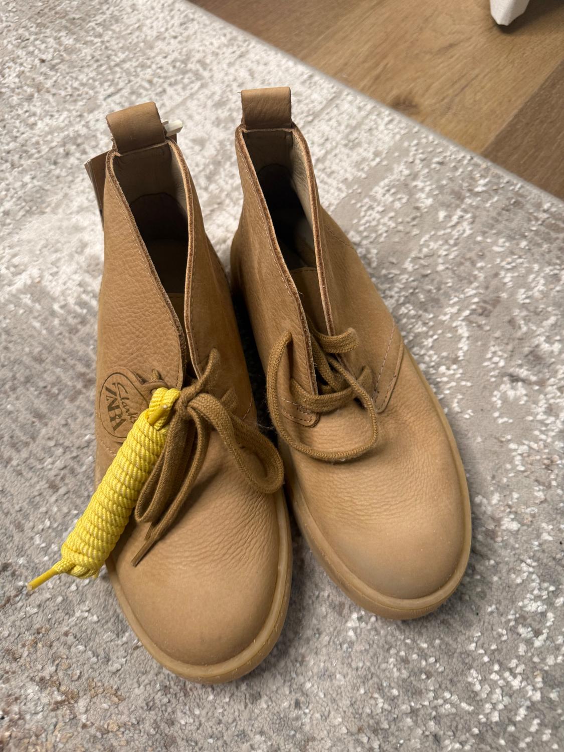 Light brown leather boots with laces and yellow detail, on a gray rug and wooden floor. Visible text: "ZNA".
