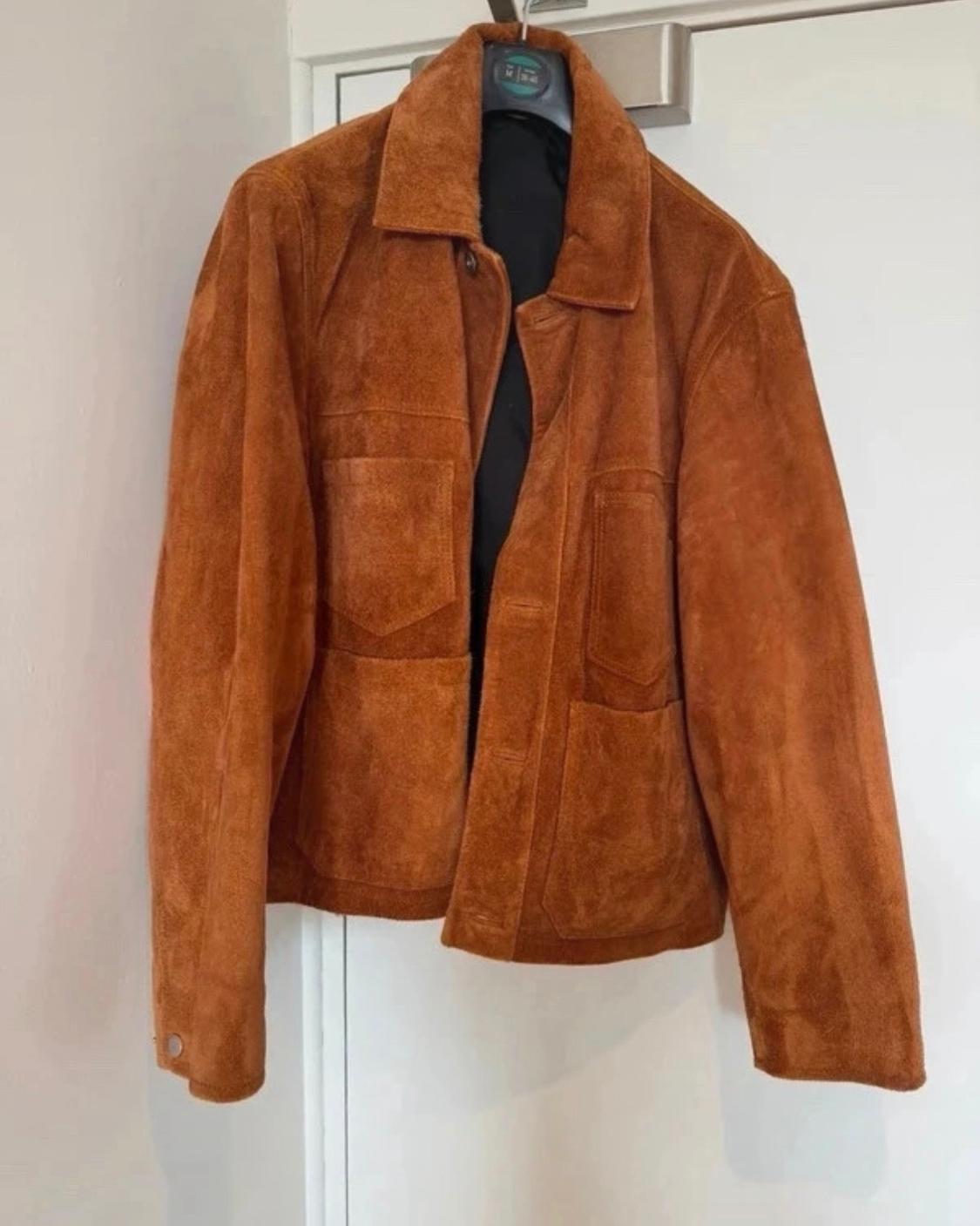 Brown suede jacket with a collar and patch pockets, hanging on a hanger over a white door.