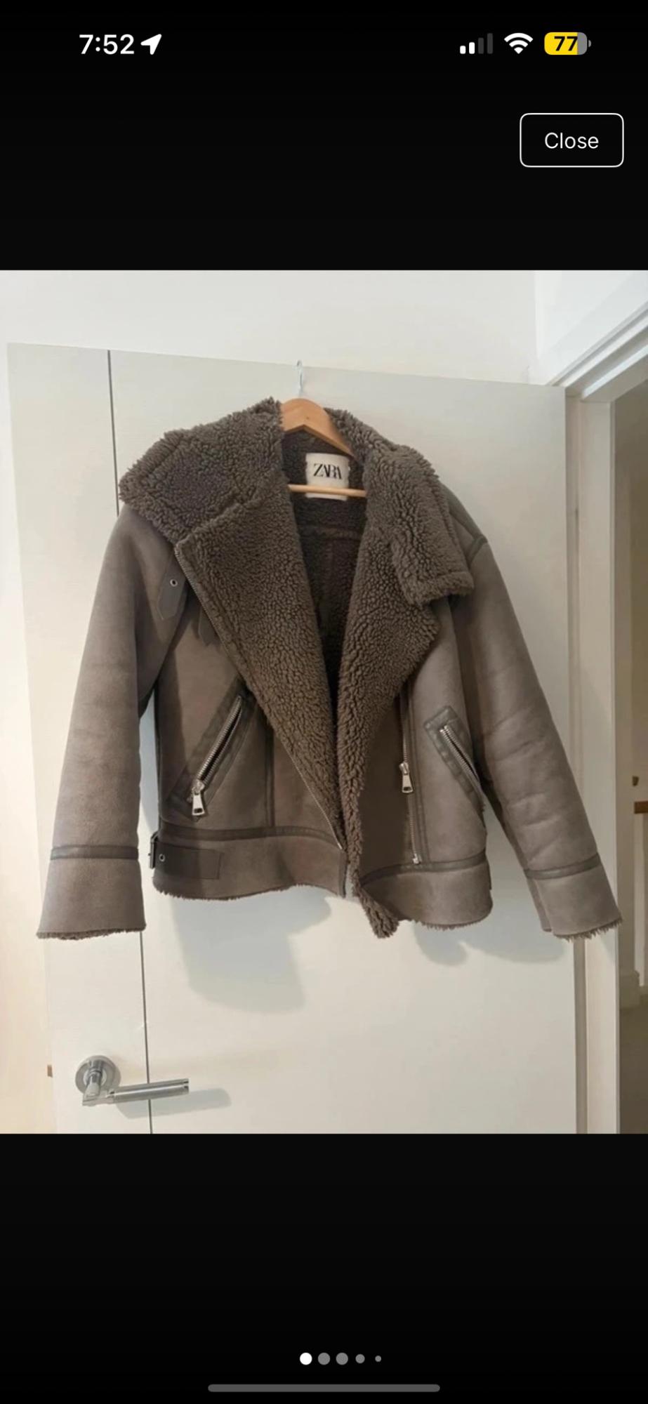 Grey jacket with a fur lining and zips, hanging from a coat hanger over a white door. Label with the text ZARA.