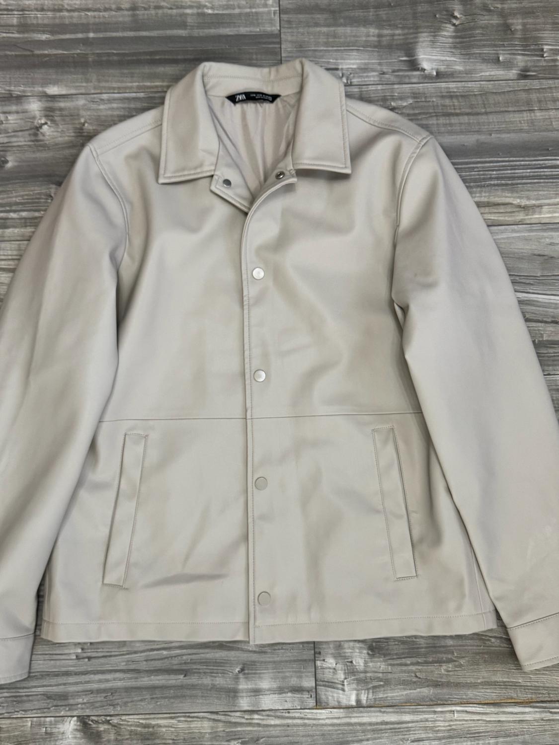 Light beige jacket with a collar, press studs and pockets, on a wooden surface. ZARA can be read on the inner label.