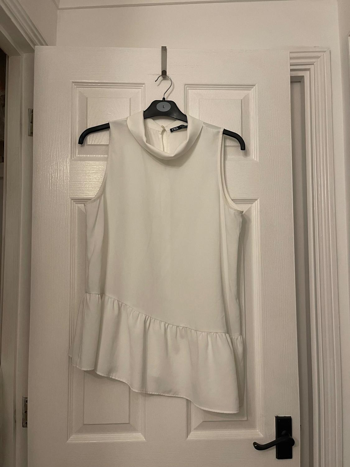 Sleeveless white high-neck blouse with a frill hem, hanging on a black hanger over a white door.