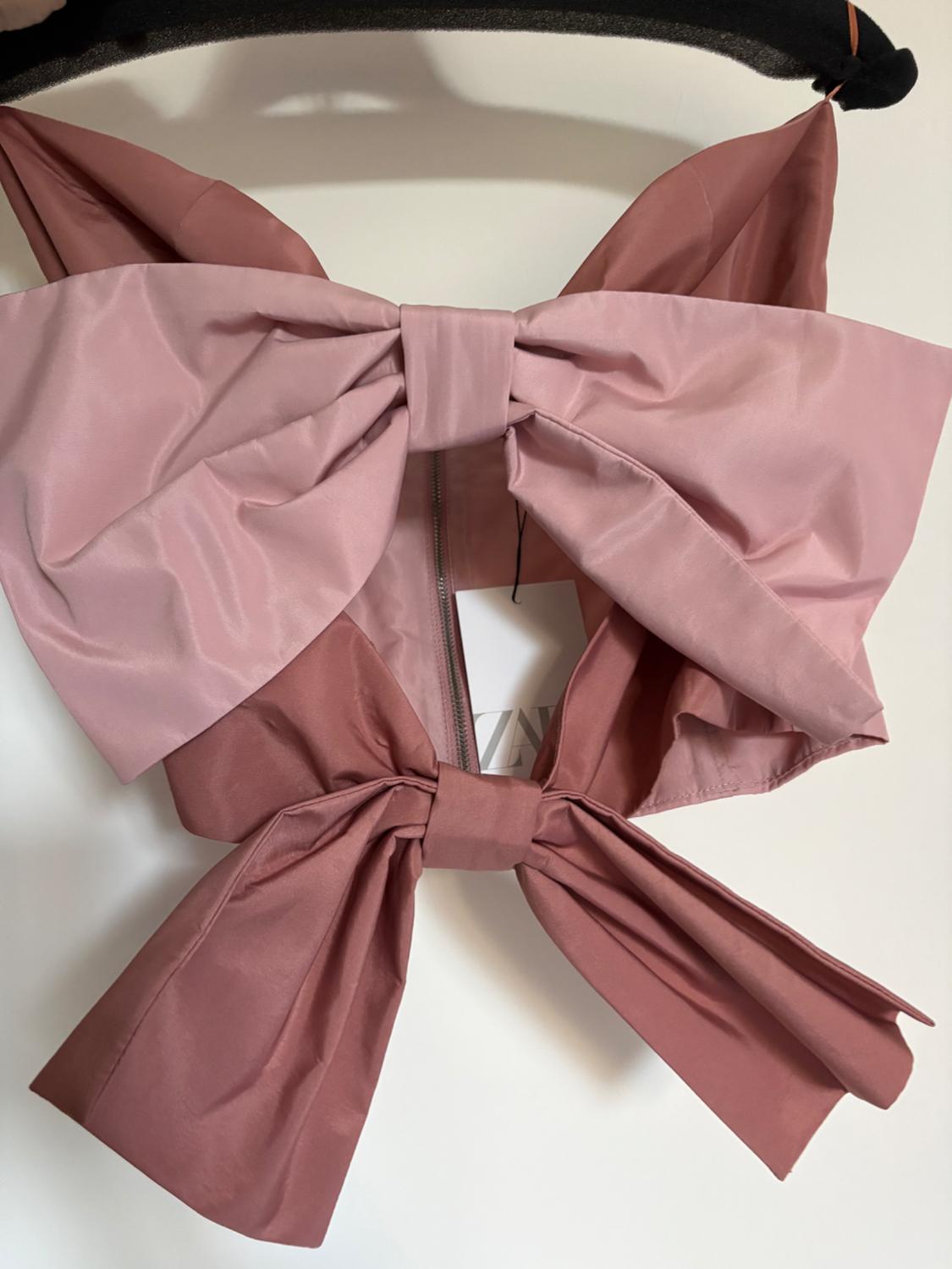 Two-tone pink camisole top hanging on a hanger. It features two large bows, a central zip and a label with the text ZA.