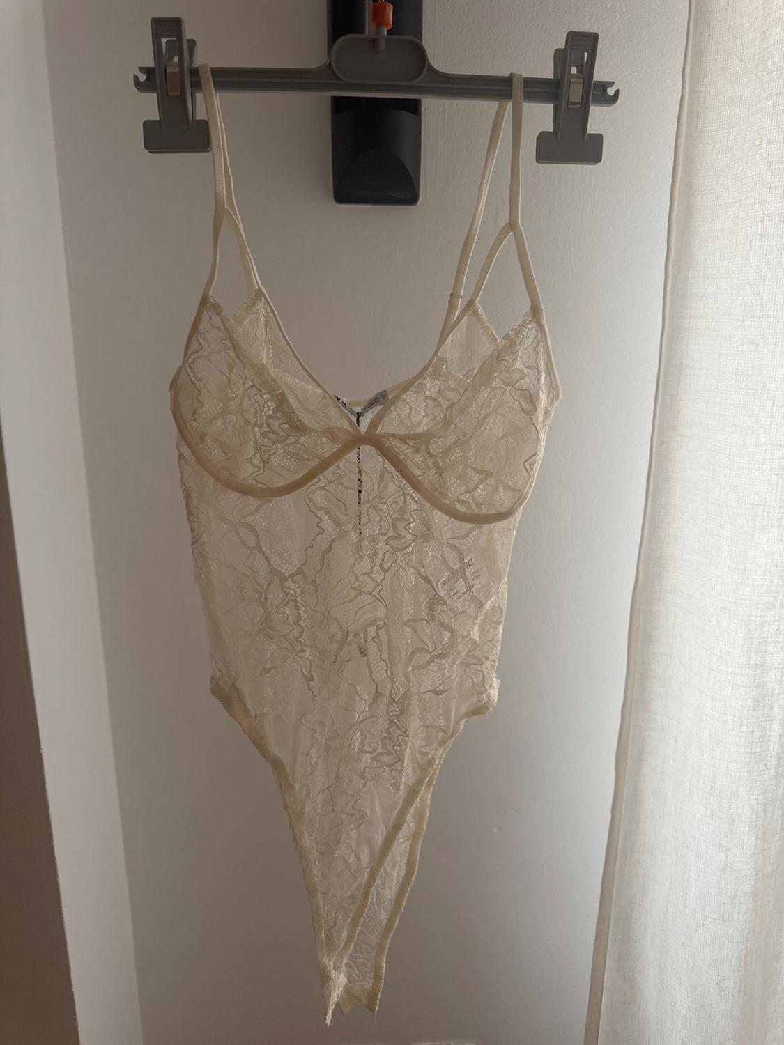 Beige lace bodysuit hanging on a grey coat hanger against a white wall, with soft light streaming in through a curtain.