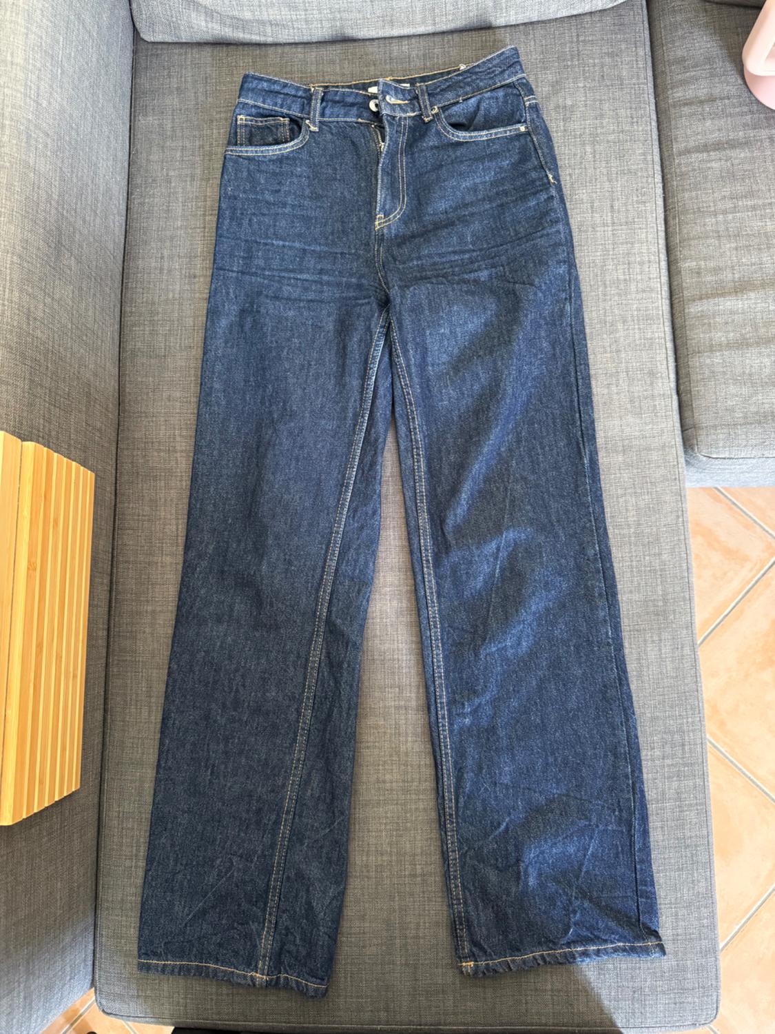 Dark blue, straight-cut denim jeans with light stitching, laid out on a grey sofa in a casual and relaxed setting.