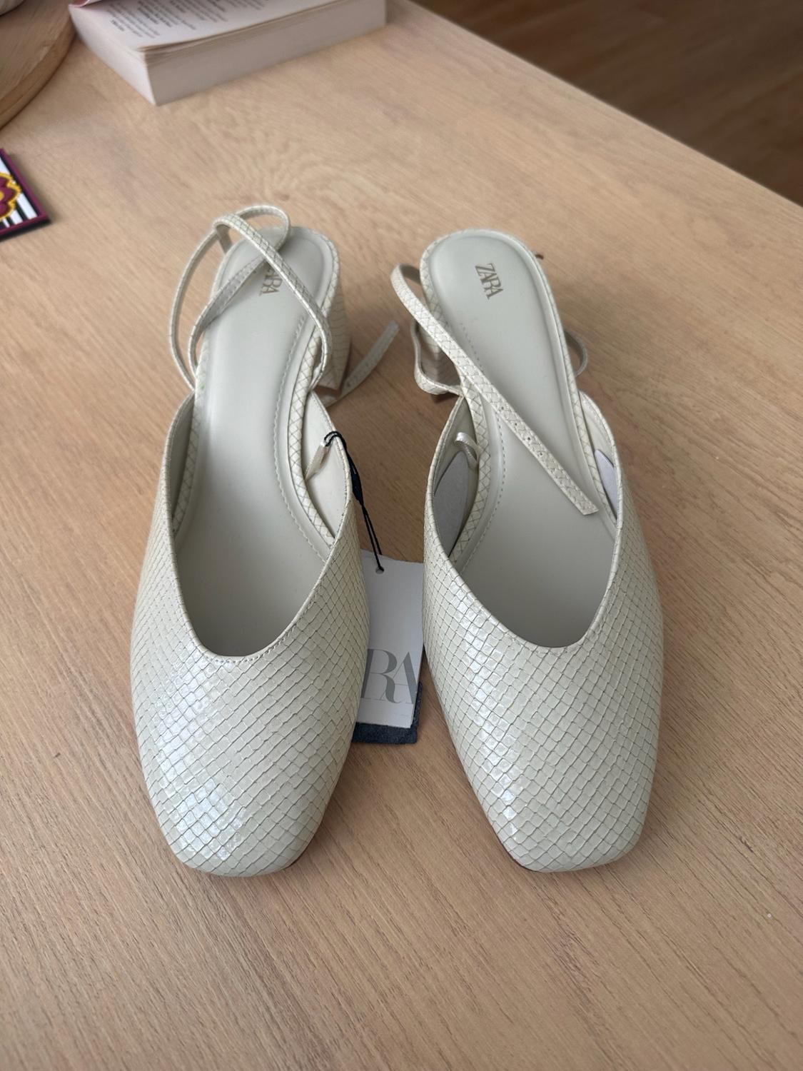 Cream-coloured textured shoes with a low heel and thin straps on a wooden table. Relaxed atmosphere. Visible text: ZARA.