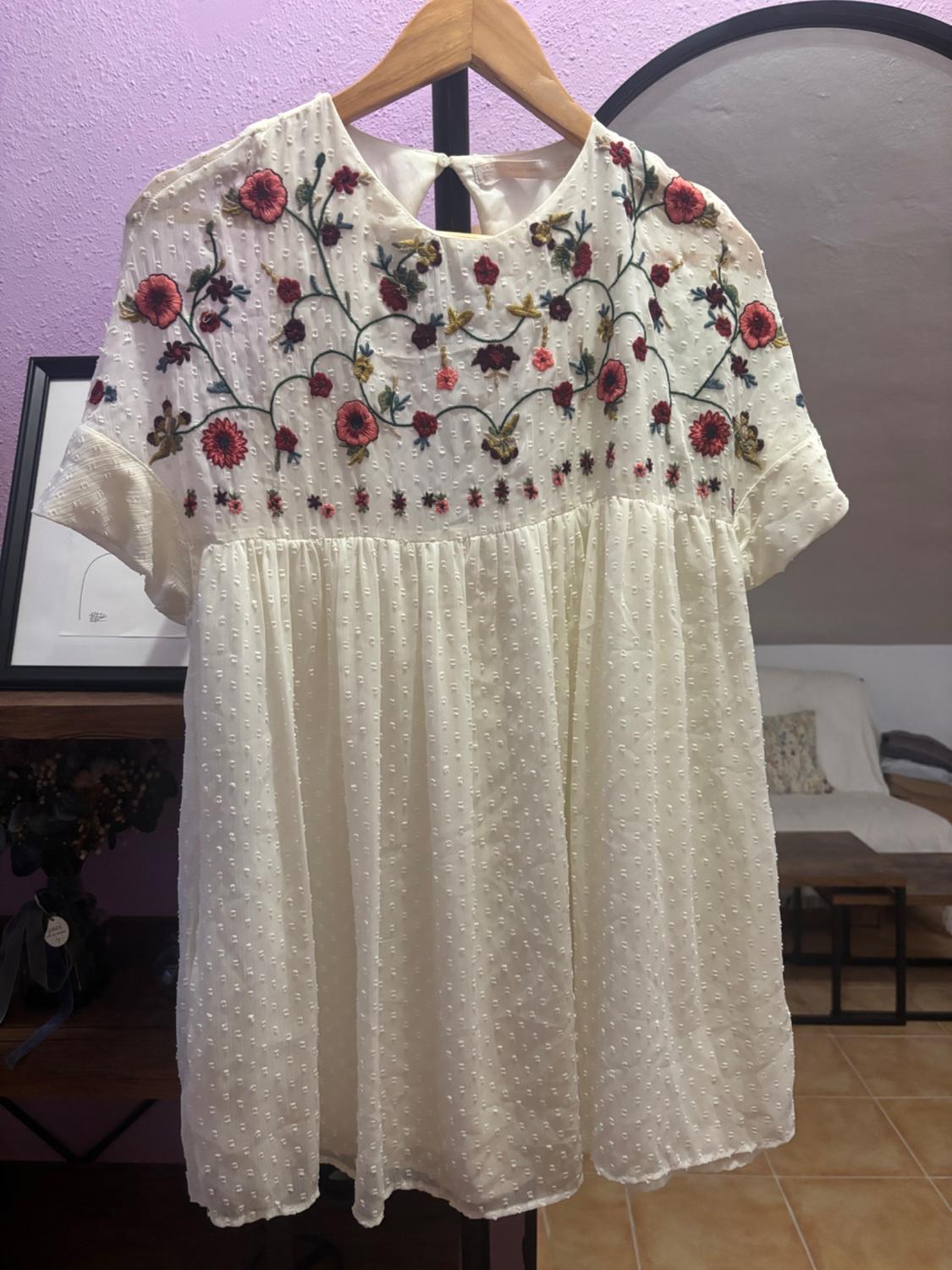 White dress with red and green floral embroidery, hanging on a wooden hanger. Background with lilac wall and mirror.